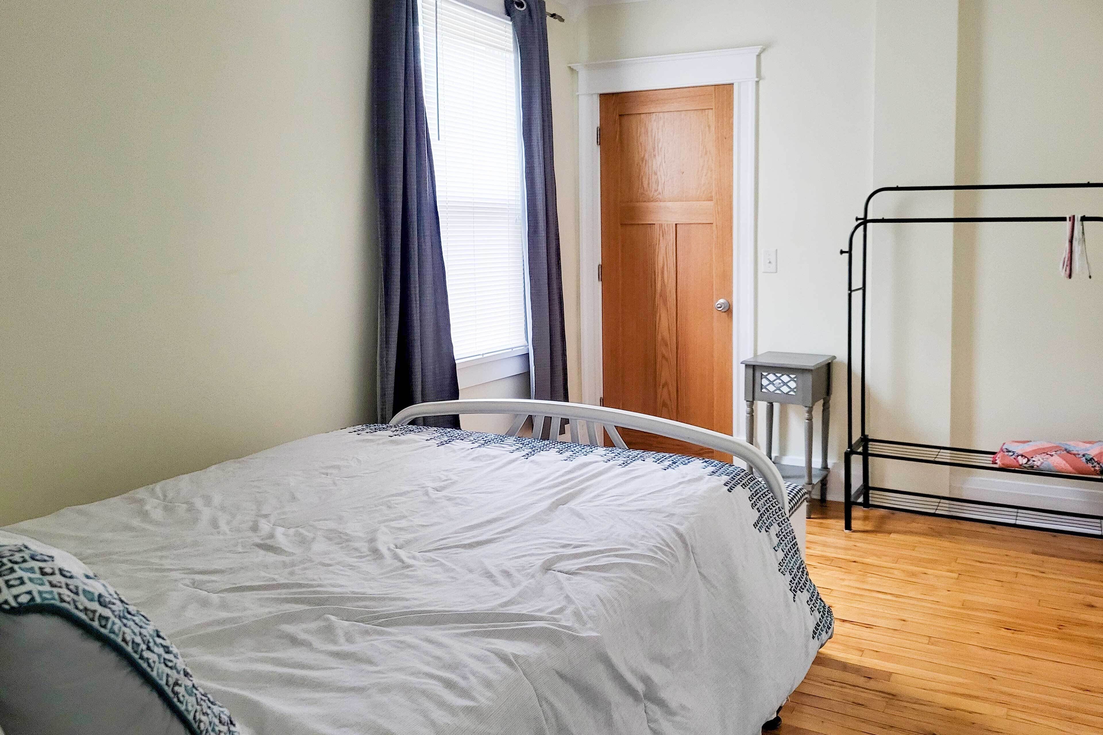 A bedroom with a bed, a wooden door, a window with curtains, a metal rack, and a small table.
