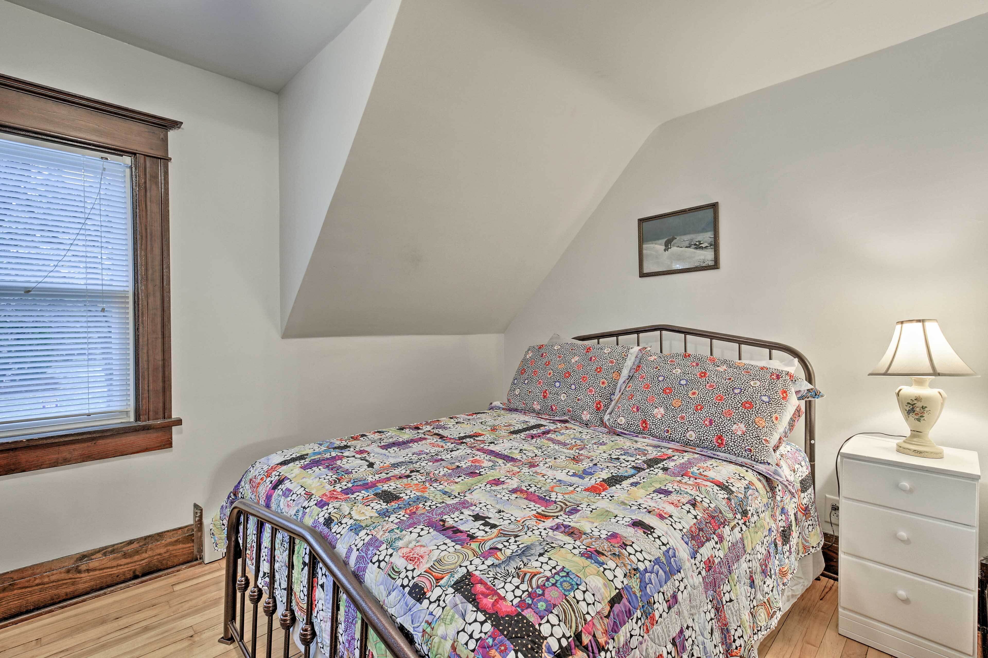 Quarto com cama, mesa de cabeceira, abajur, janela com persianas e um quadro na parede.