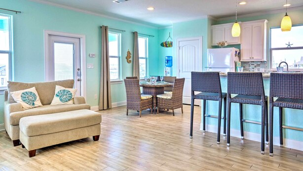 A modern kitchen with white cabinets, a dining area with wicker chairs, and a beige sofa with decorative pillows. - Furnished Deck + Ocean Views: Surfside Beach Home (Freeport)