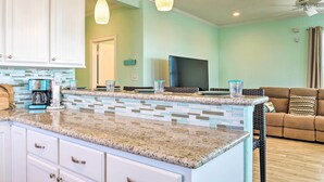 A kitchen with a granite countertop, white cabinets, and a large island. There is a flat-screen TV mounted on the wall, a wicker sofa, and a ceiling fan. - Furnished Deck + Ocean Views: Surfside Beach Home (Freeport)