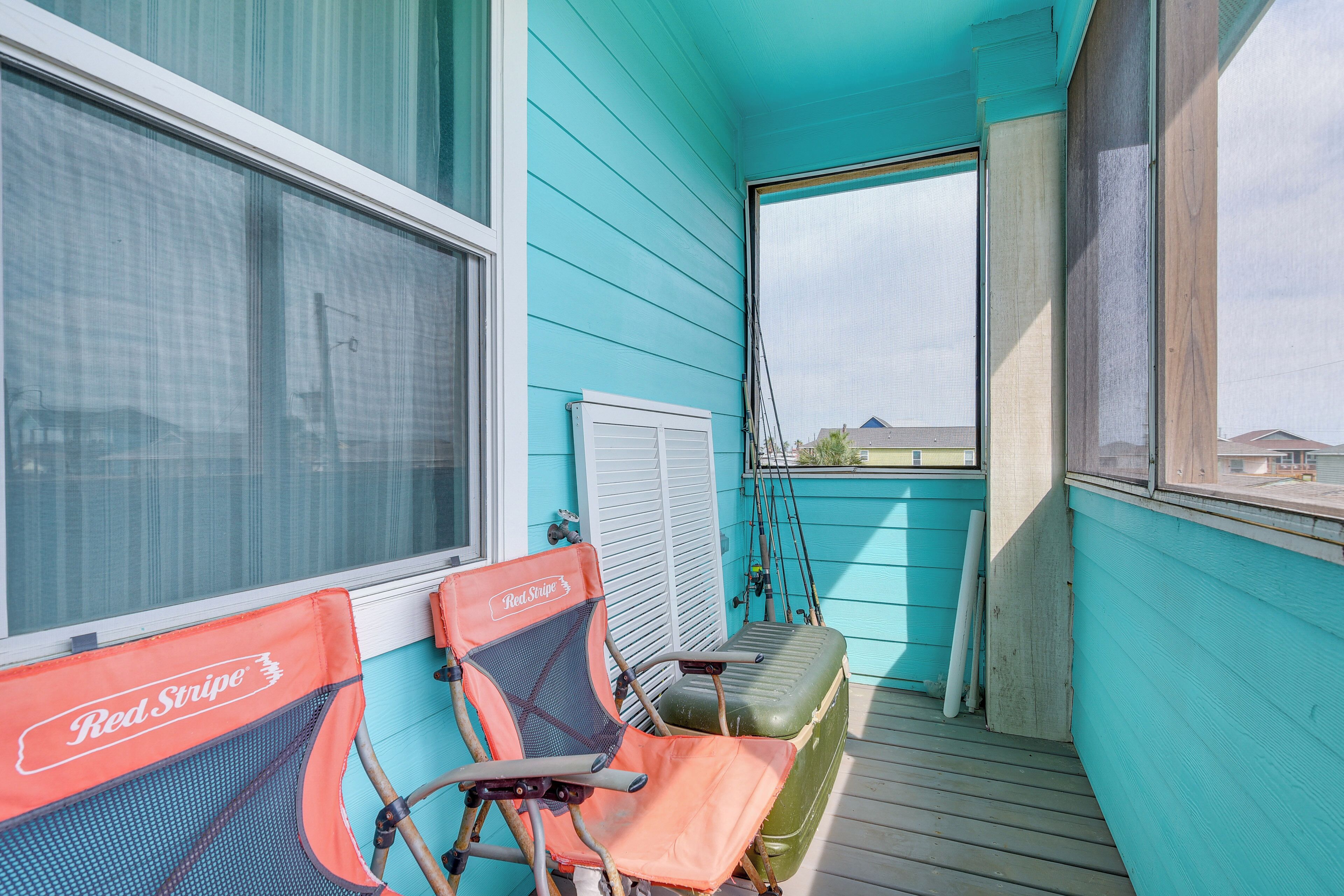 Un espace extérieur abrité, avec des murs turquoise, une chaise rouge et noire, une valise verte et des cannes à pêche appuyées contre le mur.