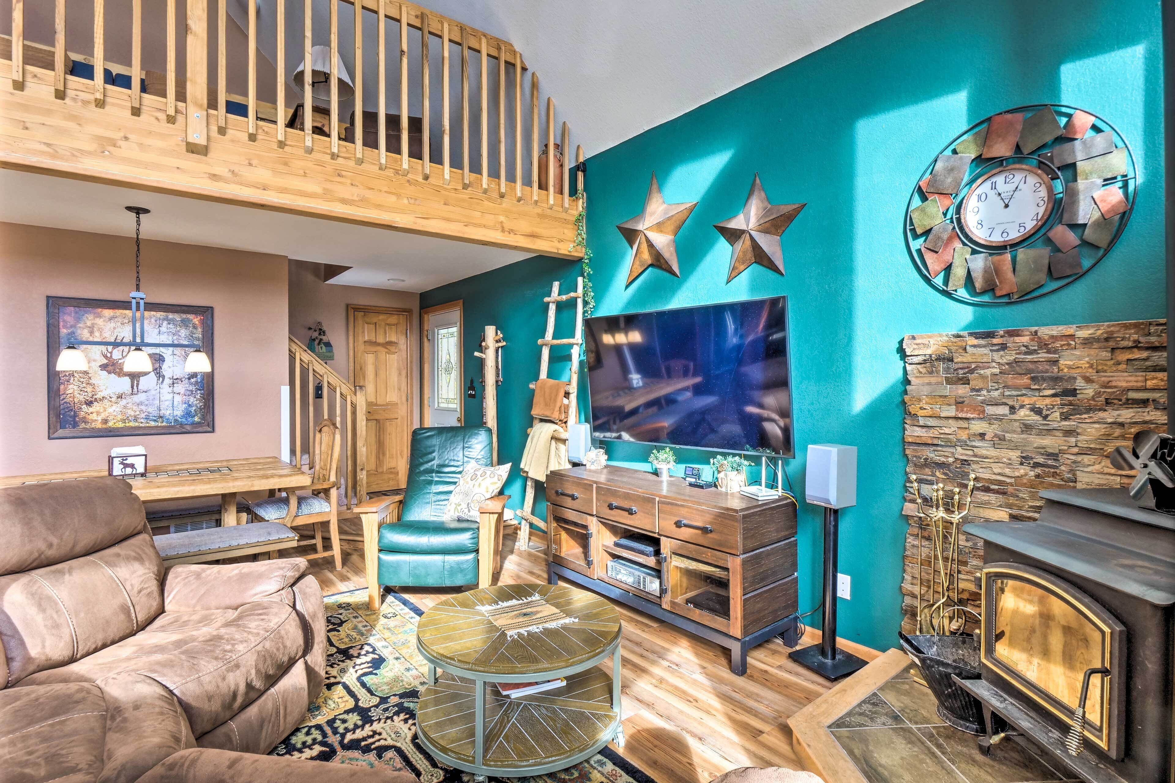 A spacious living room with a high ceiling, a wooden staircase, a large flat-screen TV, a fireplace, and a cozy seating area.
