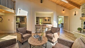 A spacious living room with a high ceiling and wooden beams, furnished with leather sofas and a round coffee table. A kitchen and dining area are visible in the background. - Hot Tub, Pool Table: Luxe Reno Chalet w/ Mtn View! (Reno)