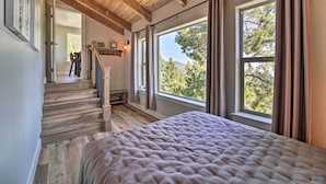 A bedroom with a large bed, wooden staircase, and large windows offering a view of trees. - Hot Tub, Pool Table: Luxe Reno Chalet w/ Mtn View! (Reno)