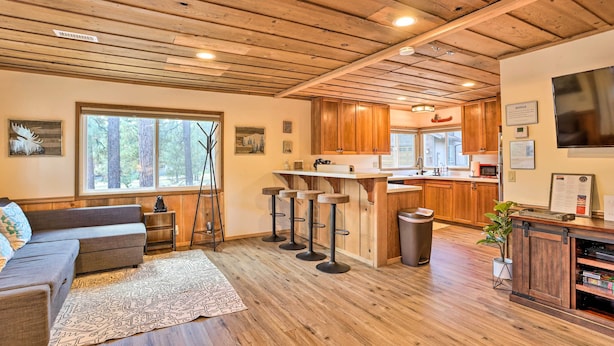 A spacious living area with a wooden ceiling, a large window, a sofa, and a bar area with stools. - Walk to Big Bear Lake: Cozy Cabin w/ Deck (Big Bear Lake)