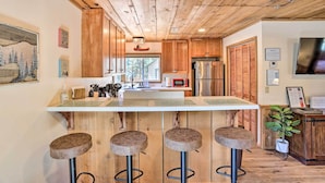 A kitchen with wooden cabinets, a stainless steel refrigerator, a microwave, and a bar area with stools. - Walk to Big Bear Lake: Cozy Cabin w/ Deck (Big Bear Lake)
