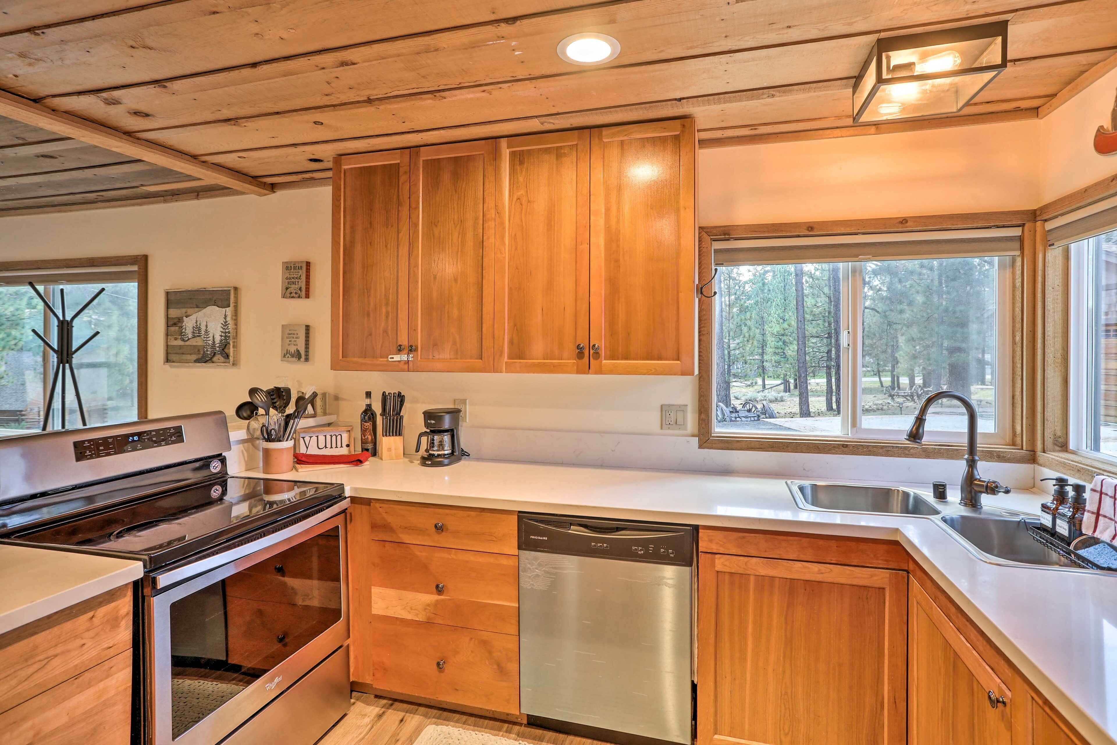 A modern kitchen with wooden cabinets, stainless steel appliances, and a large window providing a view of the forest.