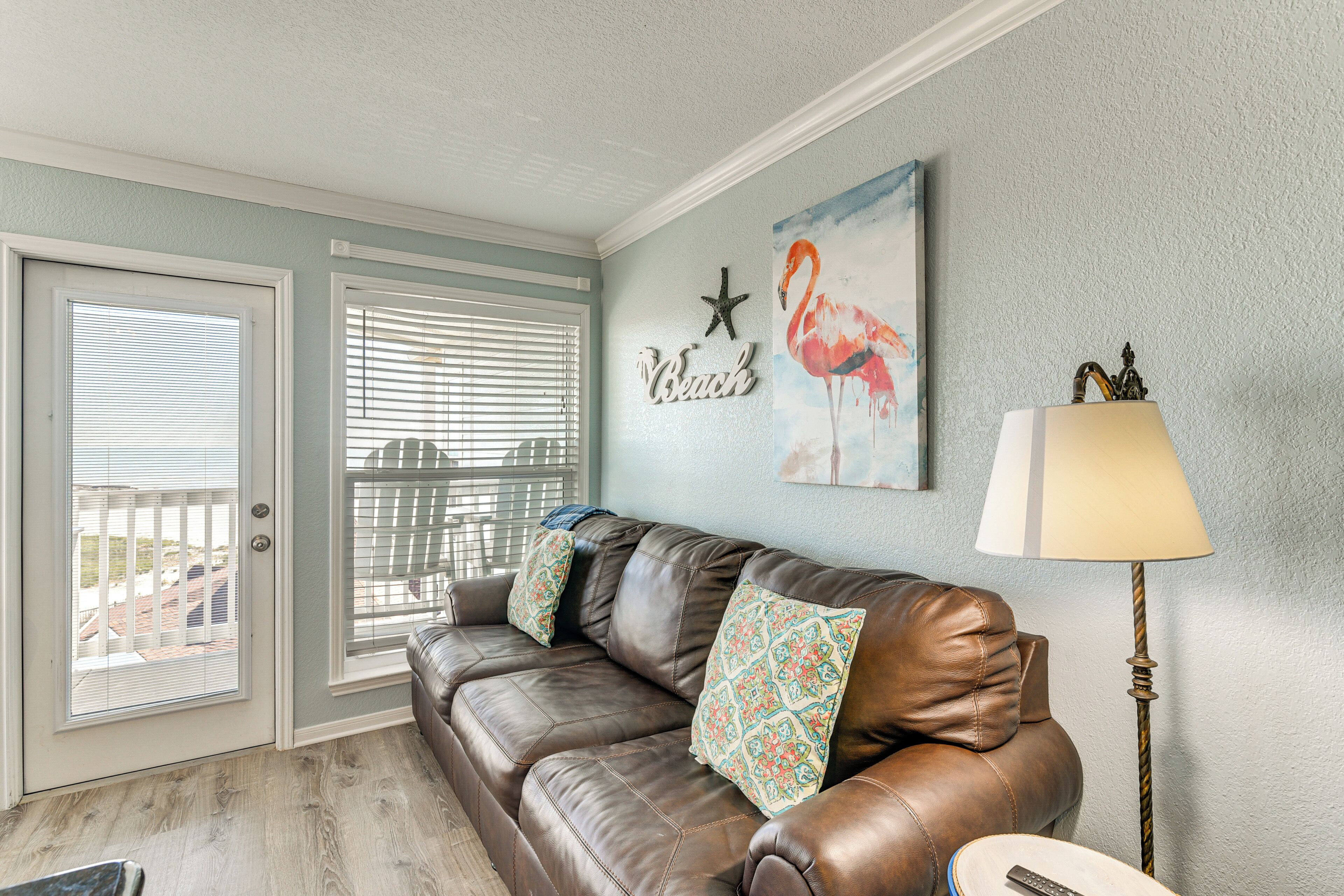 A living room with a brown leather sofa, patterned cushions, a starfish wall decoration, a flamingo painting, and a lamp.
