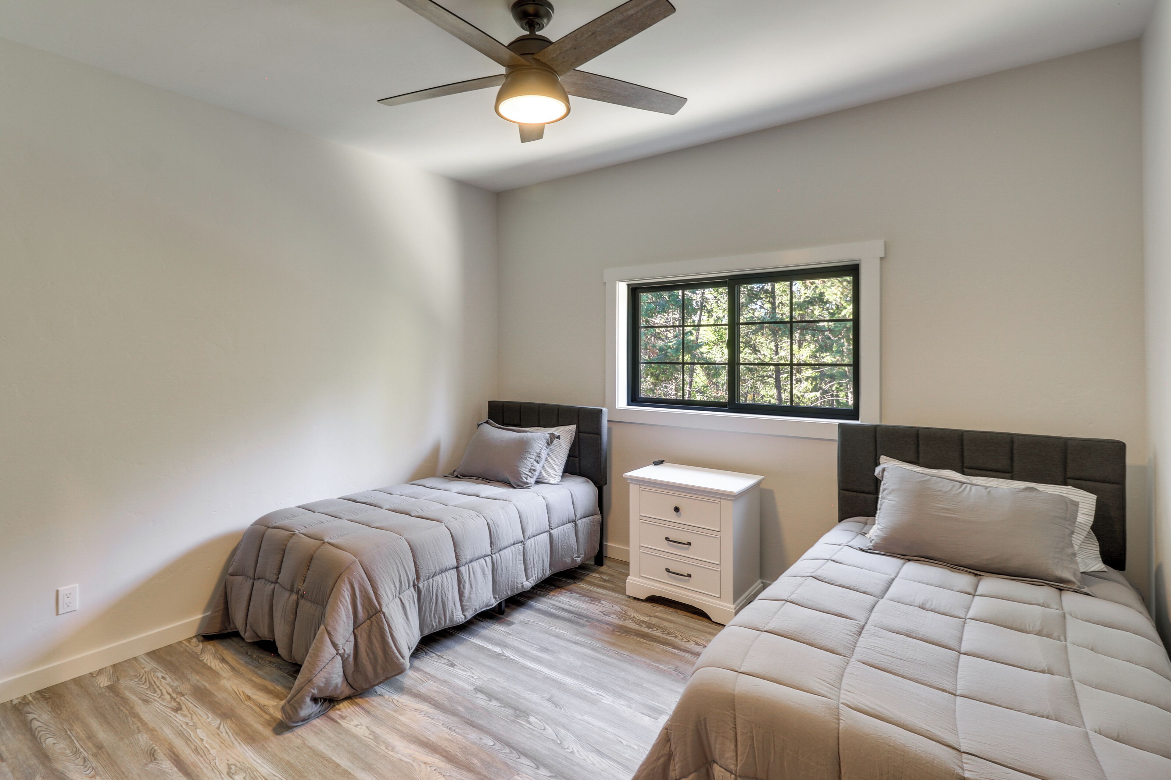 Une chambre avec deux lits, une fenêtre donnant sur la verdure, un ventilateur de plafond et une table de chevet avec des tiroirs.