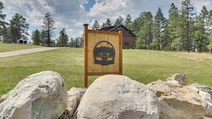 A wooden sign with a mountain and cabin design, set against a backdrop of a grassy field, trees, and a building. - Modern Kalispell Retreat: 37 Mi to Glacier NP (Kalispell)