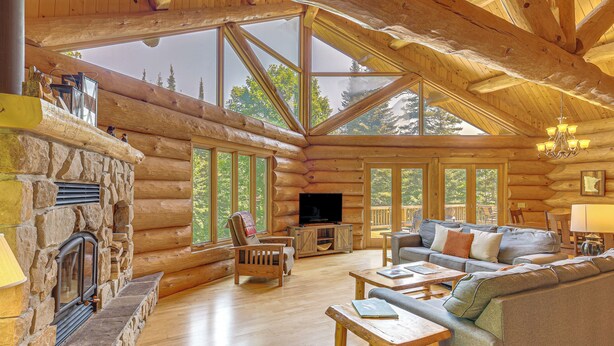 A log cabin interior with a stone fireplace, wooden furniture, large windows, and a television. - Waterfront Escape on Caribou Lake: Dock + Fire Pit (Lutsen)