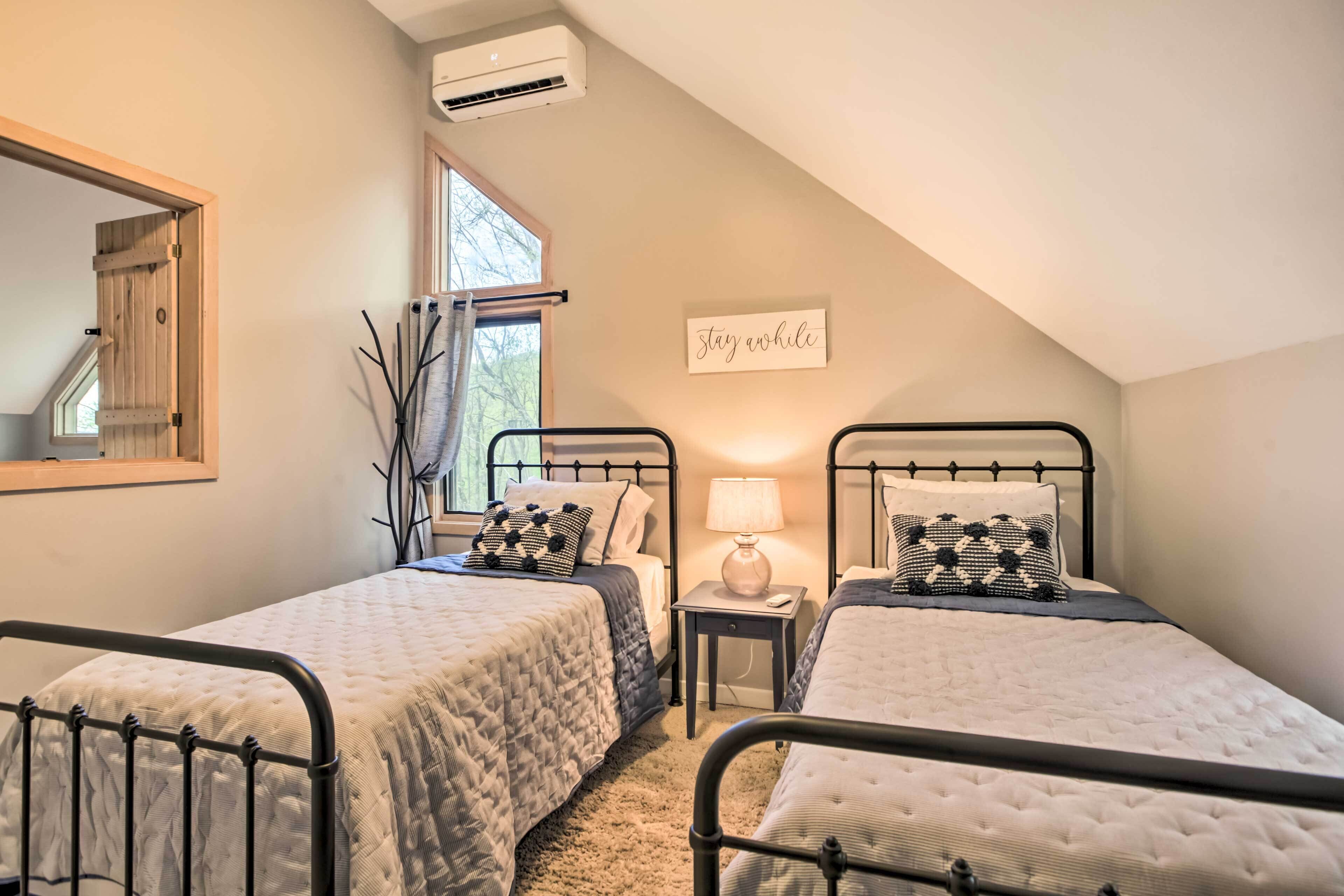 A bedroom with two beds, a window, an air conditioner, and a 'stay awhile' sign.