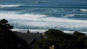 A coastal view with waves crashing onto the shore, a house with a sloped roof, and dense green trees in the foreground. - Ocean-view Lincoln City Retreat w/ 3 Viewing Decks (Lincoln City)