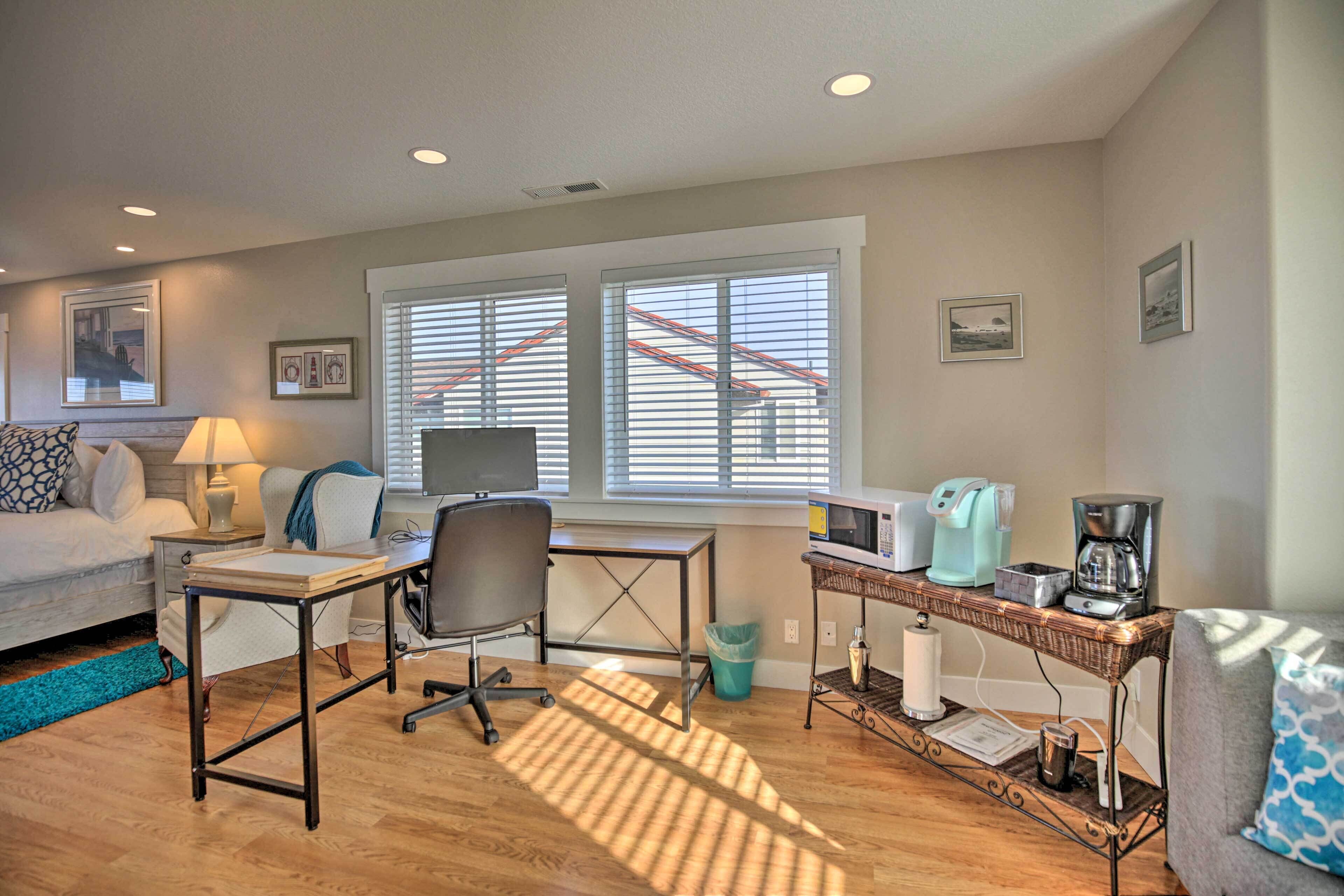 A well-lit room with a bed, desk, and seating area, featuring a window with blinds, a coffee maker, and a trash bin.