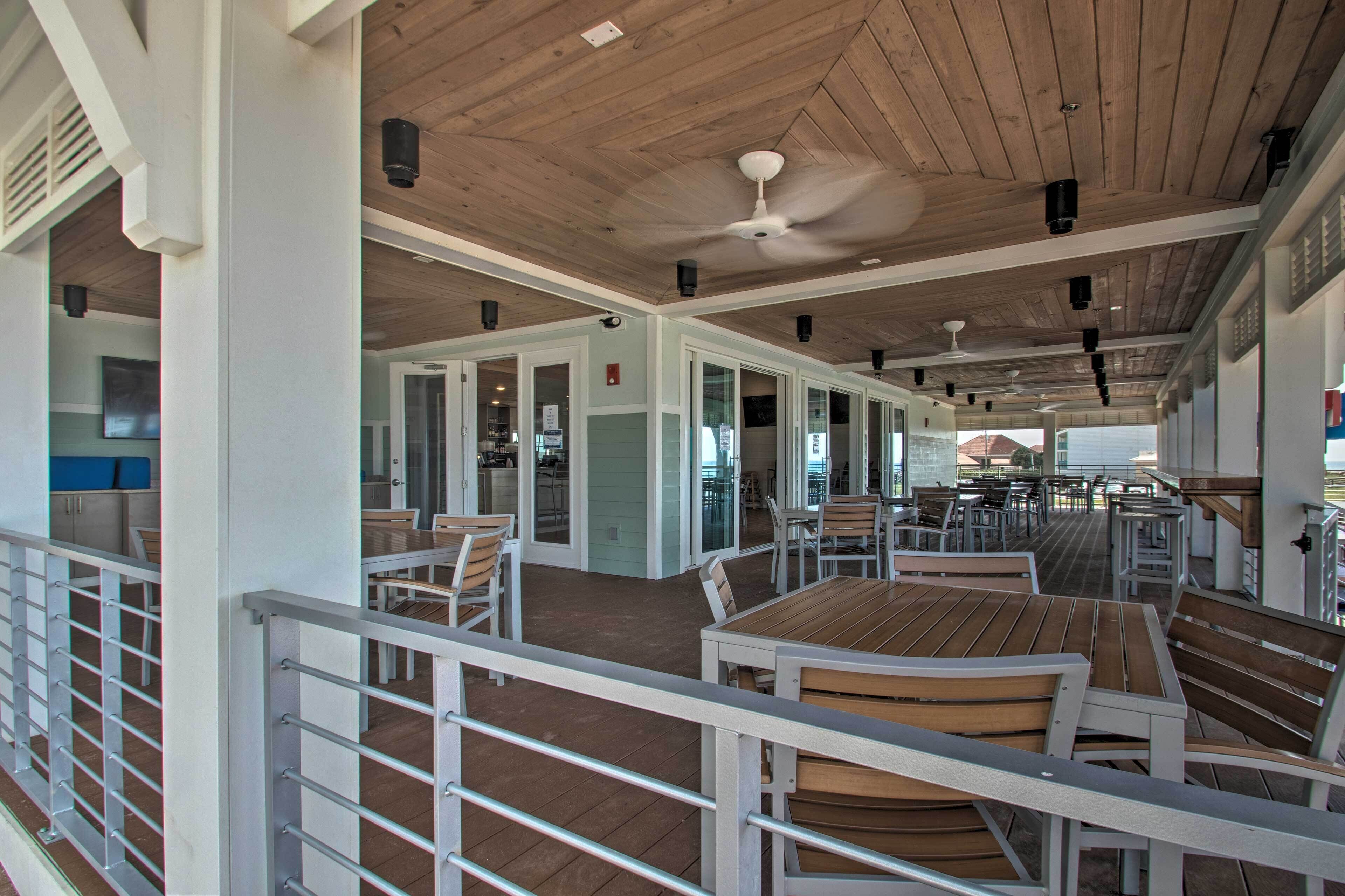 A covered outdoor dining area with wooden tables and chairs, a ceiling fan, and a view of the ocean.