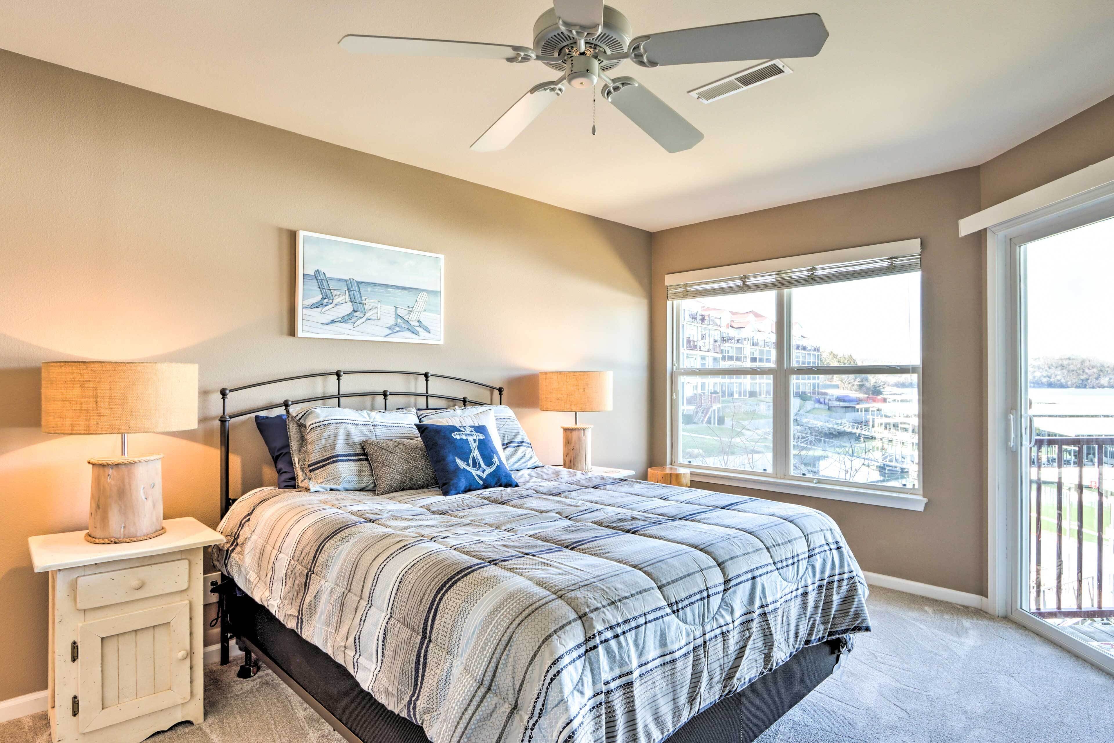 A bedroom with a bed, a ceiling fan, a window with a view, and a nightstand with a lamp.