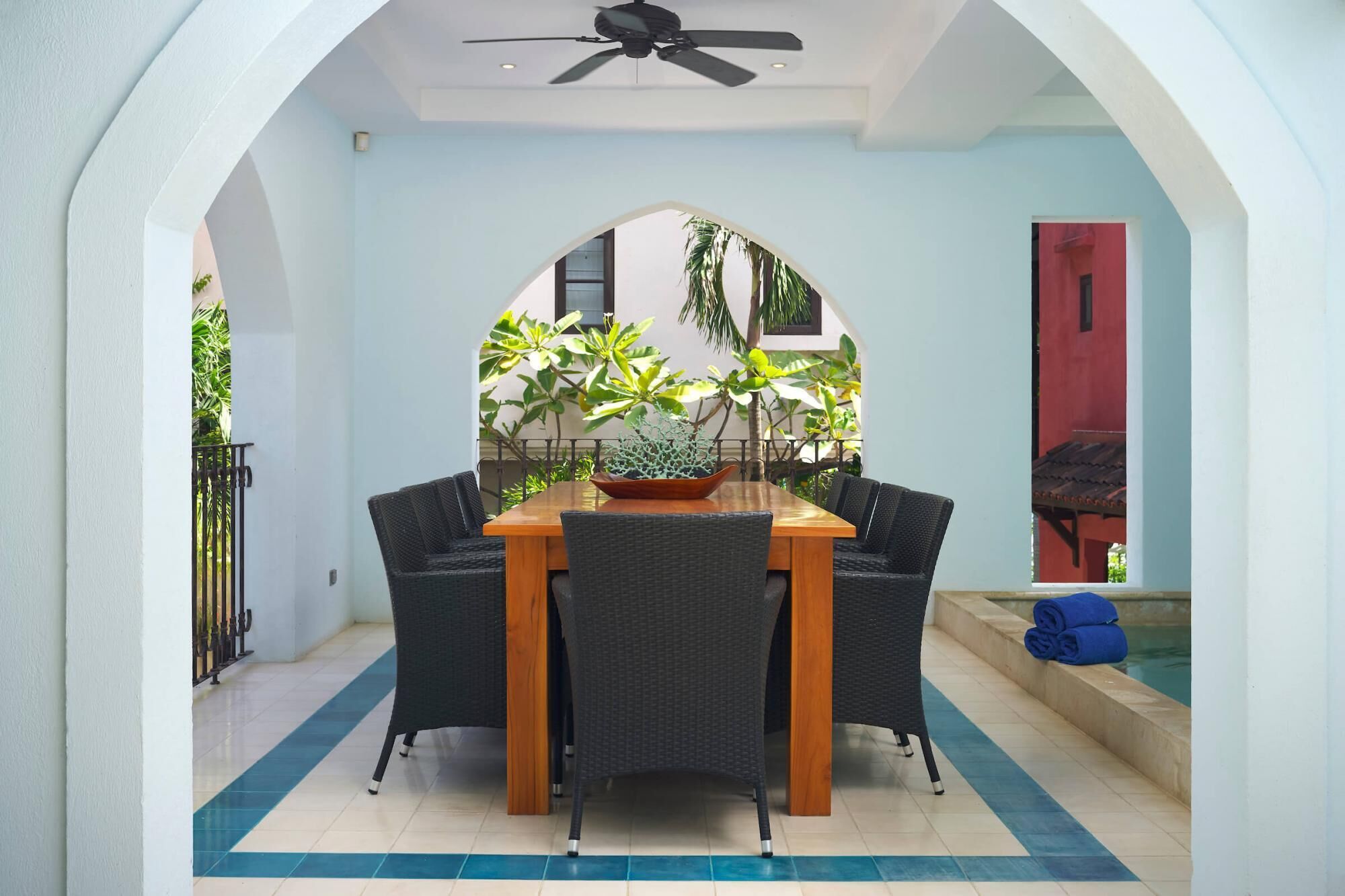 A dining area with a wooden table and wicker chairs, surrounded by a white wall with a blue-tiled floor.