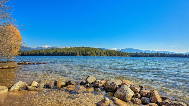 Ein klarer See mit sichtbaren Felsen im Vordergrund und einem bewaldeten Gebirge im Hintergrund.
