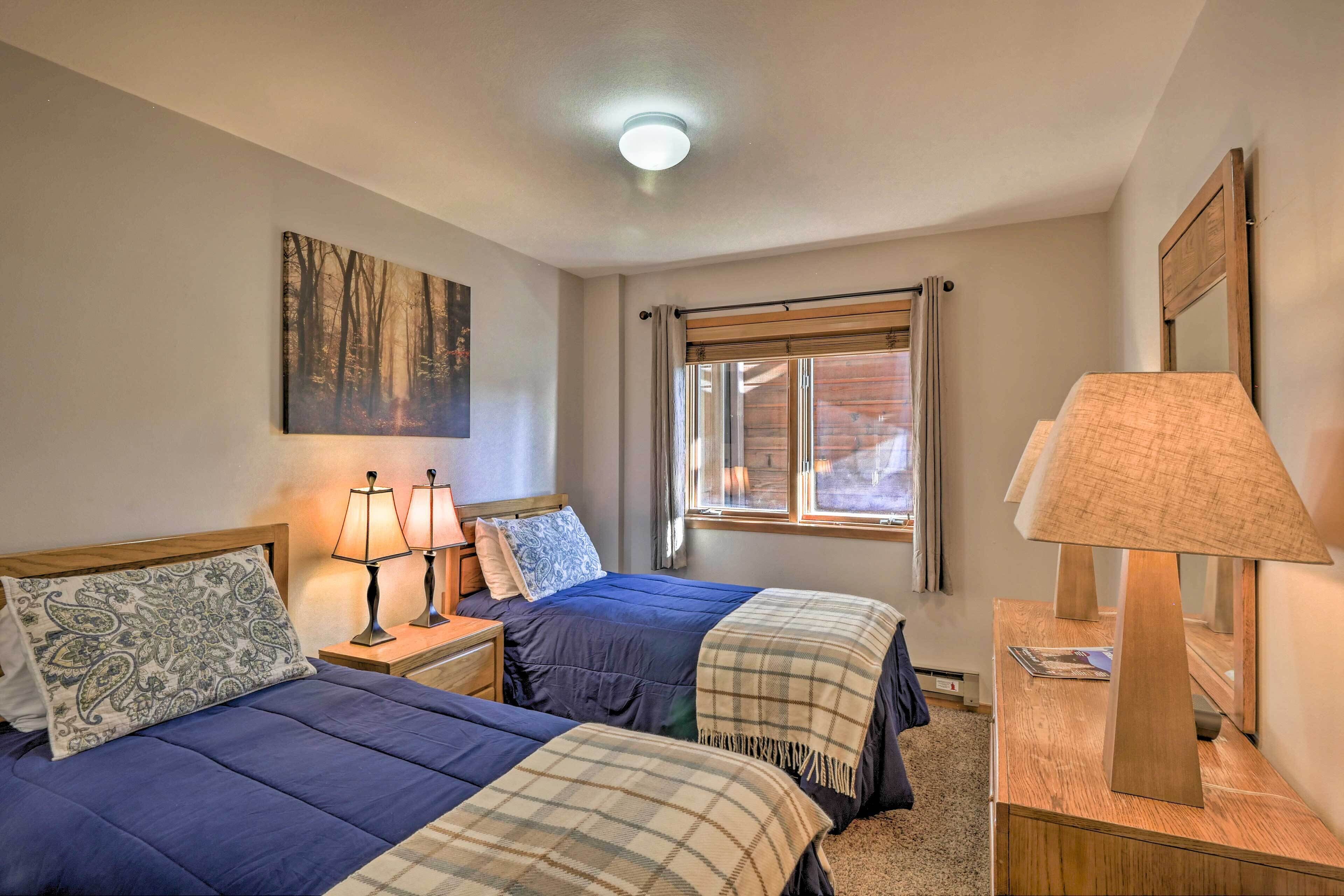 A bedroom with two beds, a nightstand with lamps, a window with curtains, and a painting on the wall.