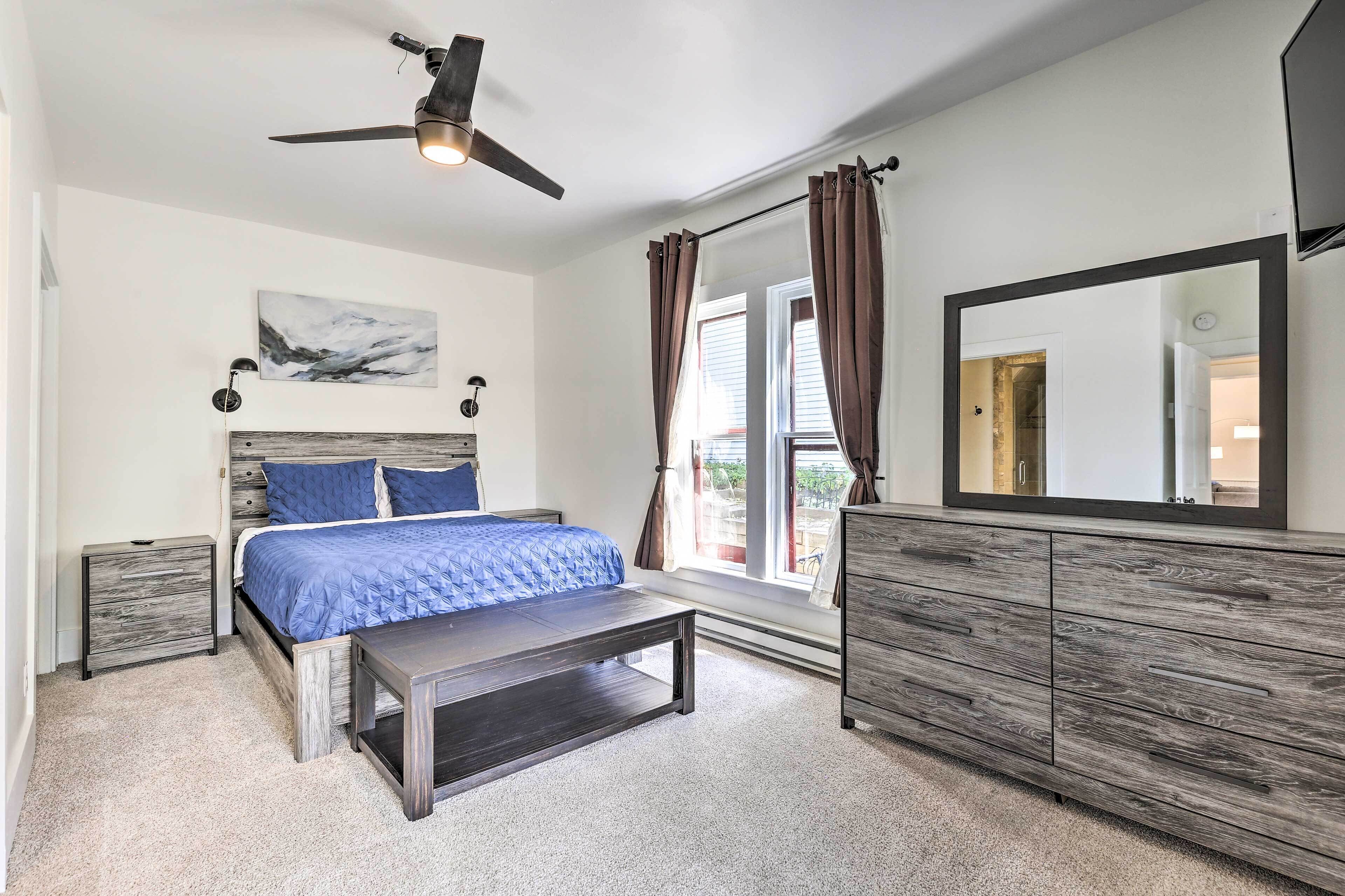 Une chambre à coucher comprenant un lit, des tables de chevet, un banc, une commode, un ventilateur de plafond, une fenêtre avec des rideaux et un miroir.