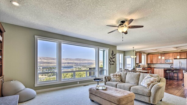 A spacious living room with a large window offering a view of a valley, a ceiling fan, and a kitchen area in the background.