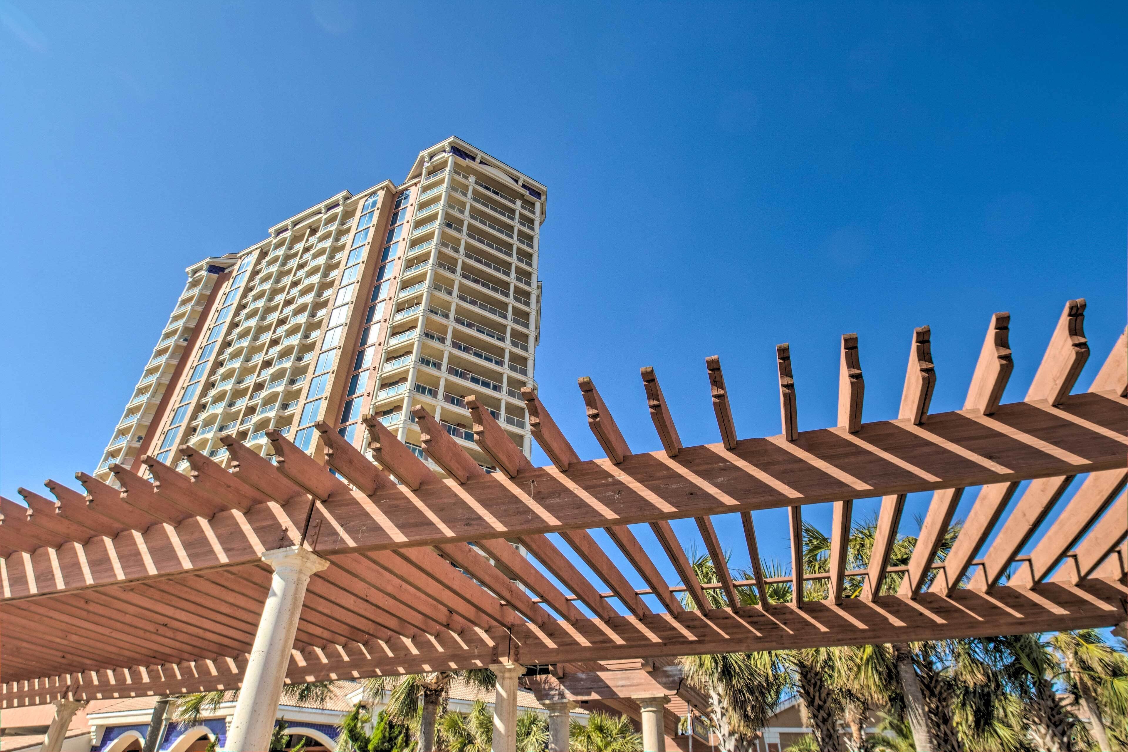 Un edificio de gran altura con varios balcones, una estructura de pérgola de madera en primer plano y palmeras al fondo.