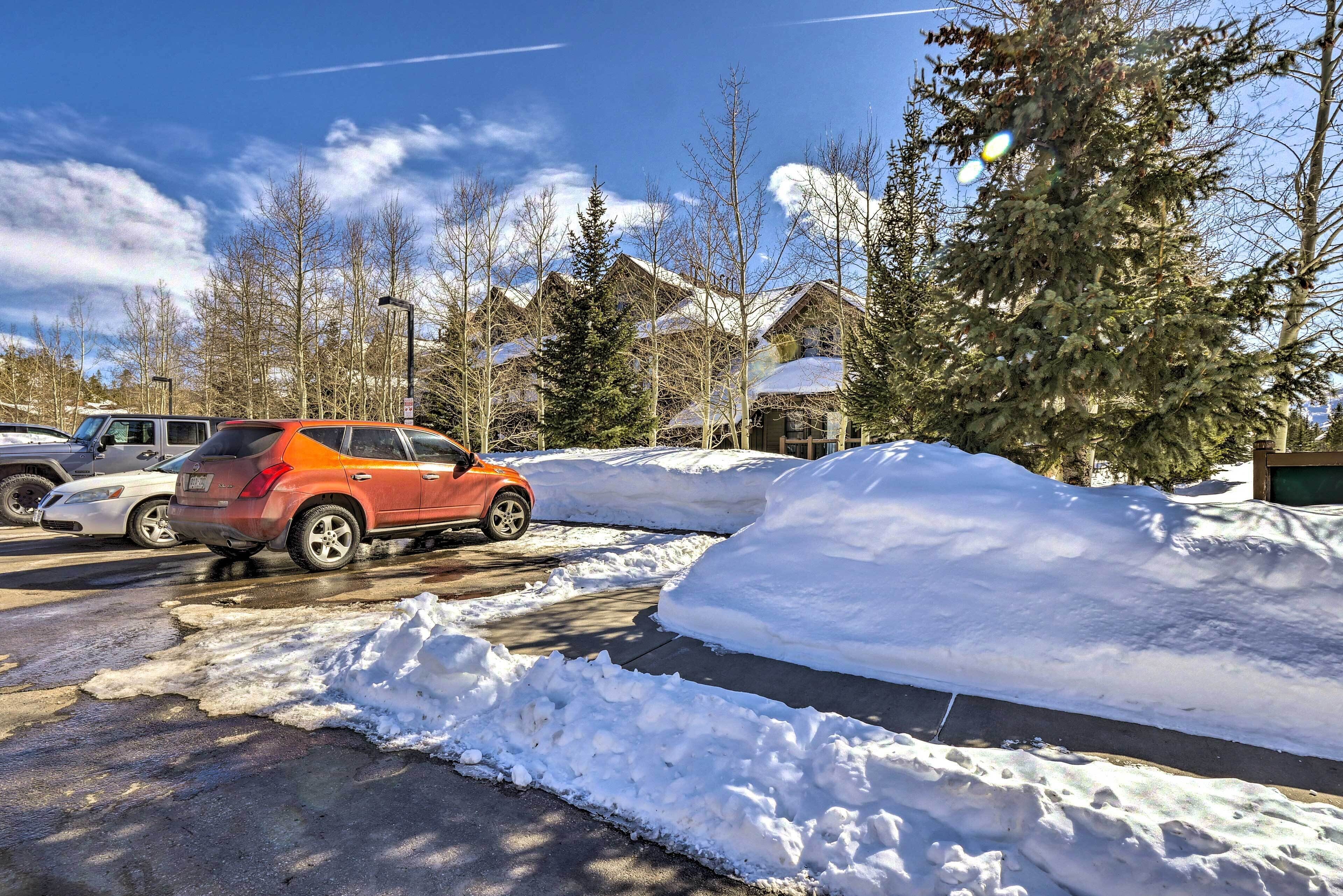 Un estacionamiento con bancos de nieve, un auto rojo y casas al fondo.