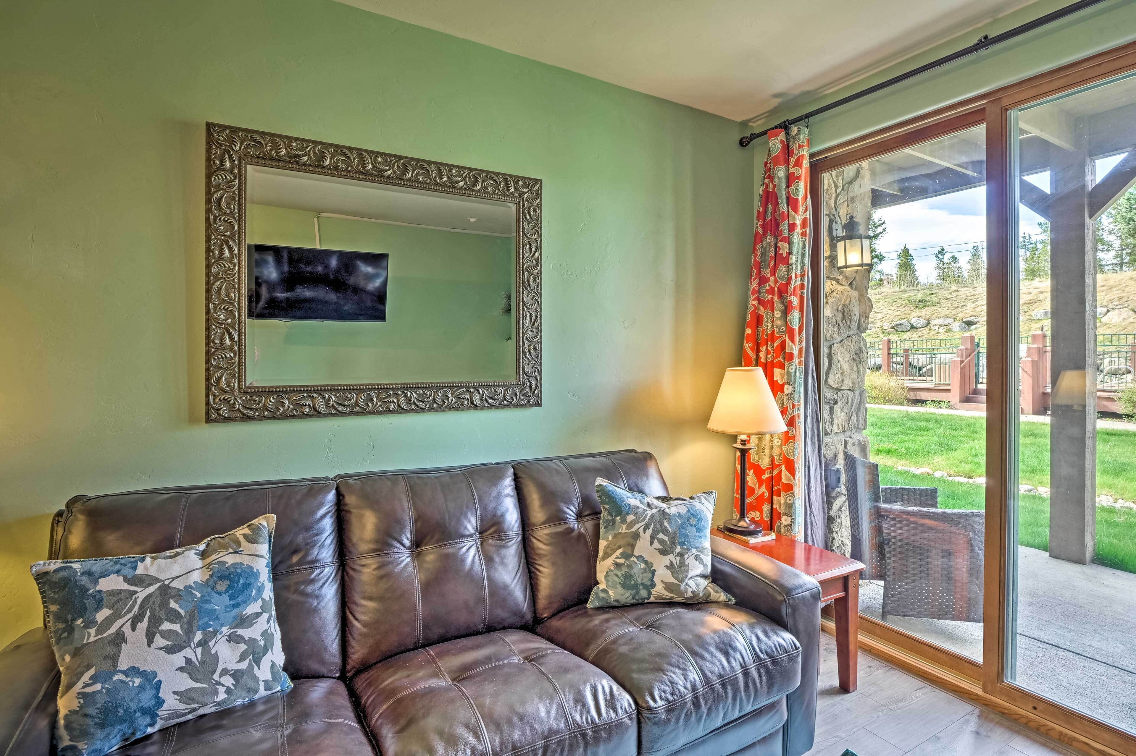 A living room with a brown leather sofa, two floral cushions, a large mirror, and a view of a garden through a glass door.
