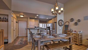 Un comedor amplio con una mesa de madera dispuesta para cuatro personas, rodeada de sillas. Un candelabro cuelga encima, y la habitación cuenta con armarios de madera y una cocina al fondo.