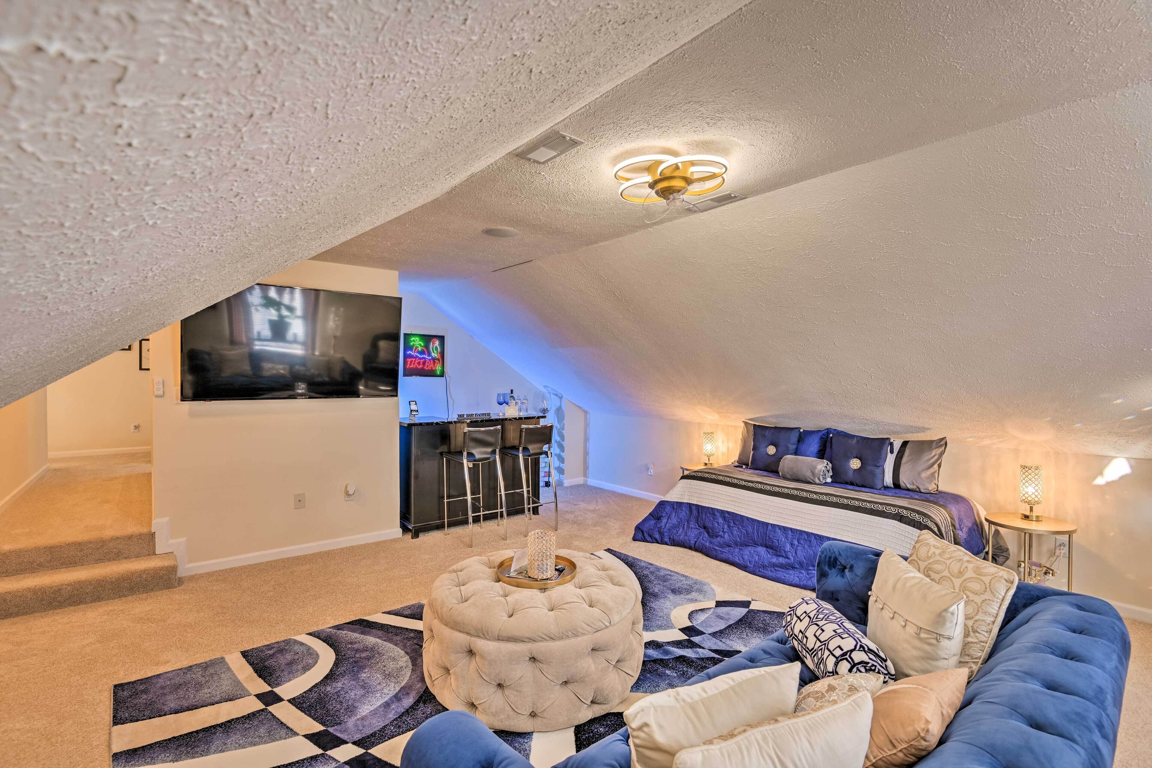 A modern loft bedroom with a large bed, a seating area with a tufted ottoman, and a kitchenette in the background.