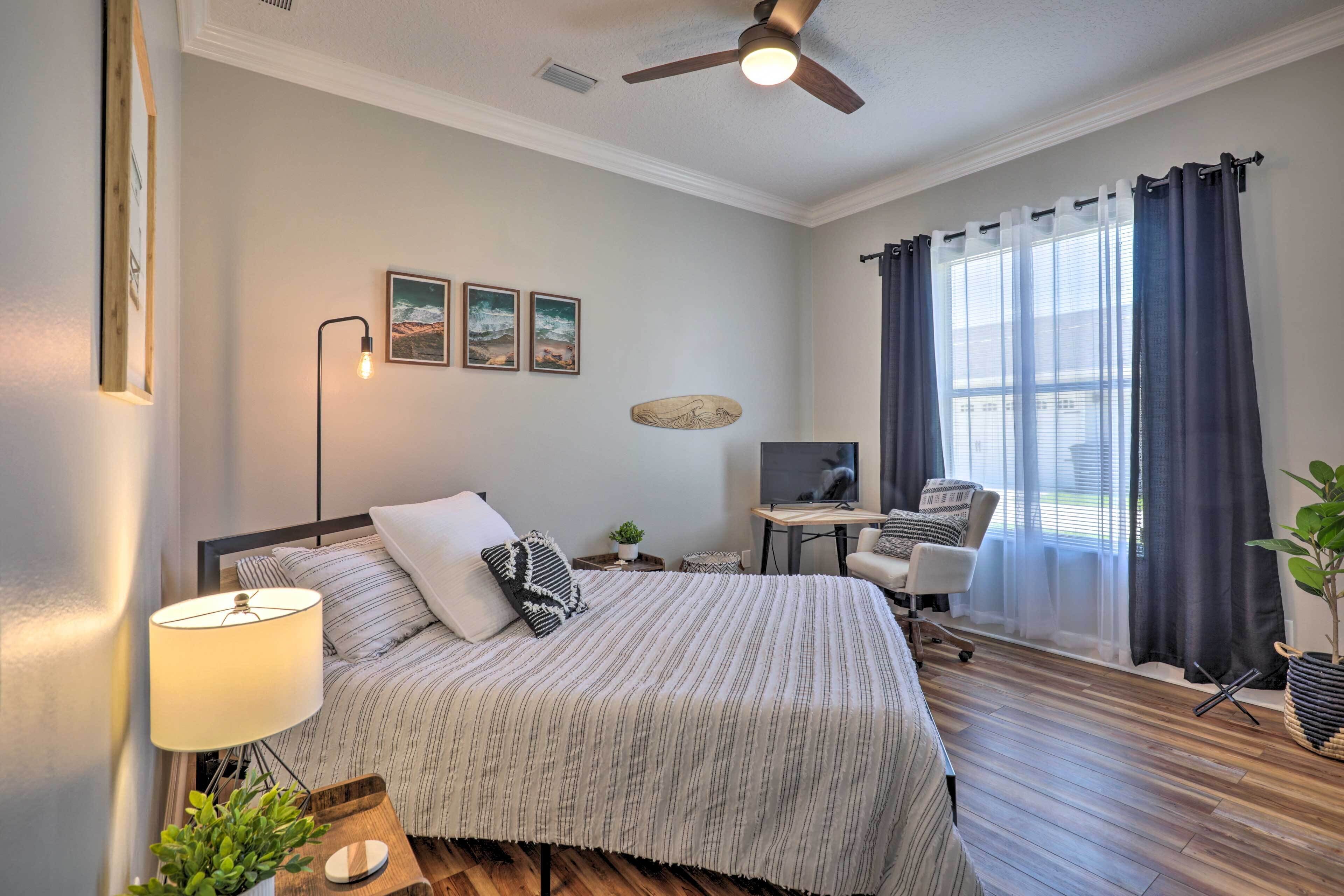 Un dormitorio con cama, escritorio, silla, ventilador de techo y ventana con cortinas.