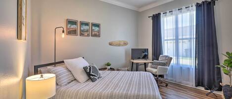 Un dormitorio con cama, escritorio, silla, ventilador de techo y ventana con cortinas.