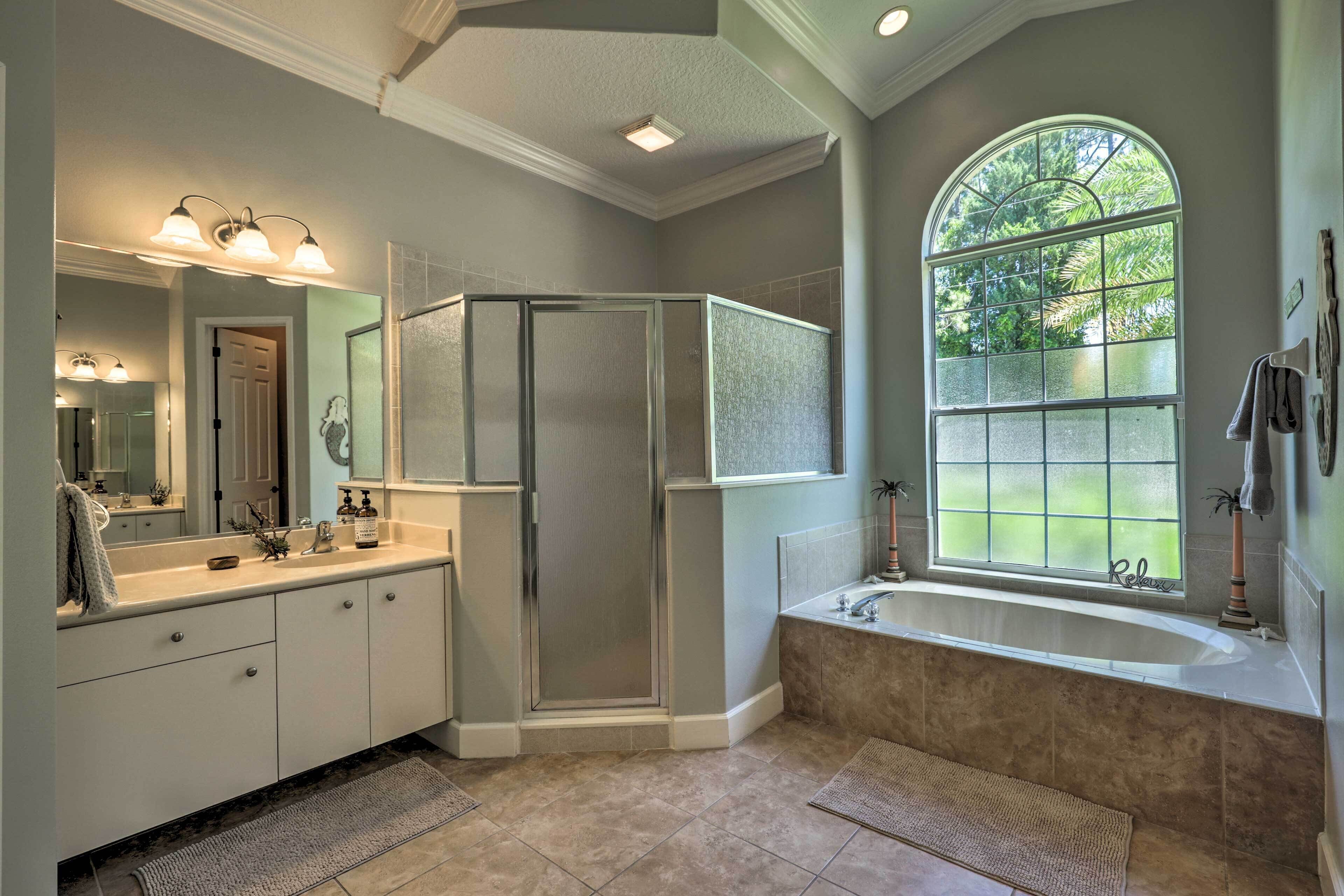 A modern bathroom with a walk-in shower, a large mirror, a bathtub, and a window with a view of trees.