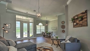 A spacious living room with a gray sectional sofa, a coffee table, and a gray armchair. There are two pendant lights, a wooden wall decor, and a dining area visible in the background. - Upscale Palm Coast Home w/ Pool: 7 Mi to Beaches! (Palm Coast)