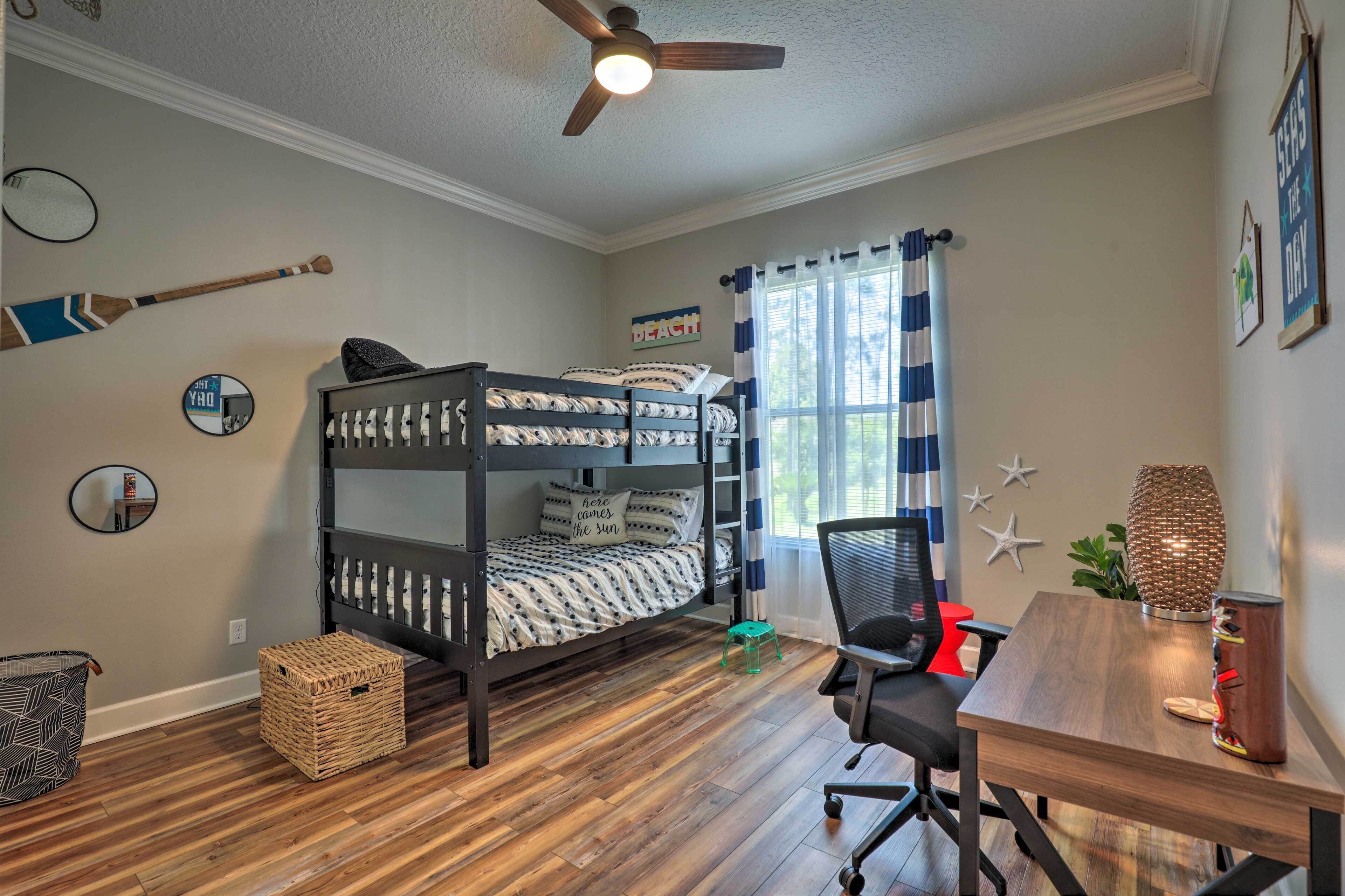 A bunk bed with a "home comes the sun" pillow, a desk with a chair, a ceiling fan, and a window with curtains.
