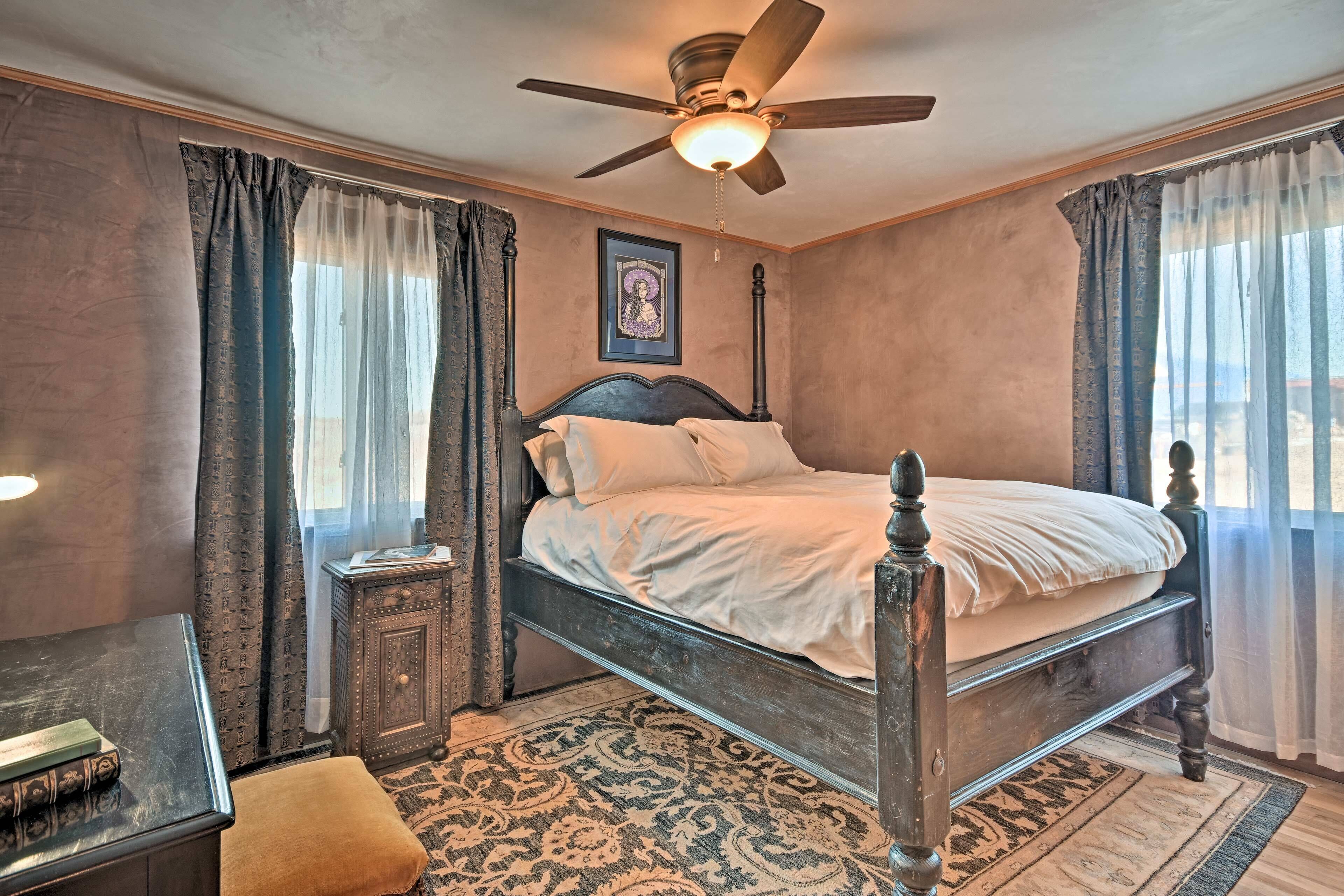 Une chambre à coucher avec un lit à baldaquin en bois, un ventilateur de plafond, une table de chevet, une petite table avec un livre et un tapis à motifs.