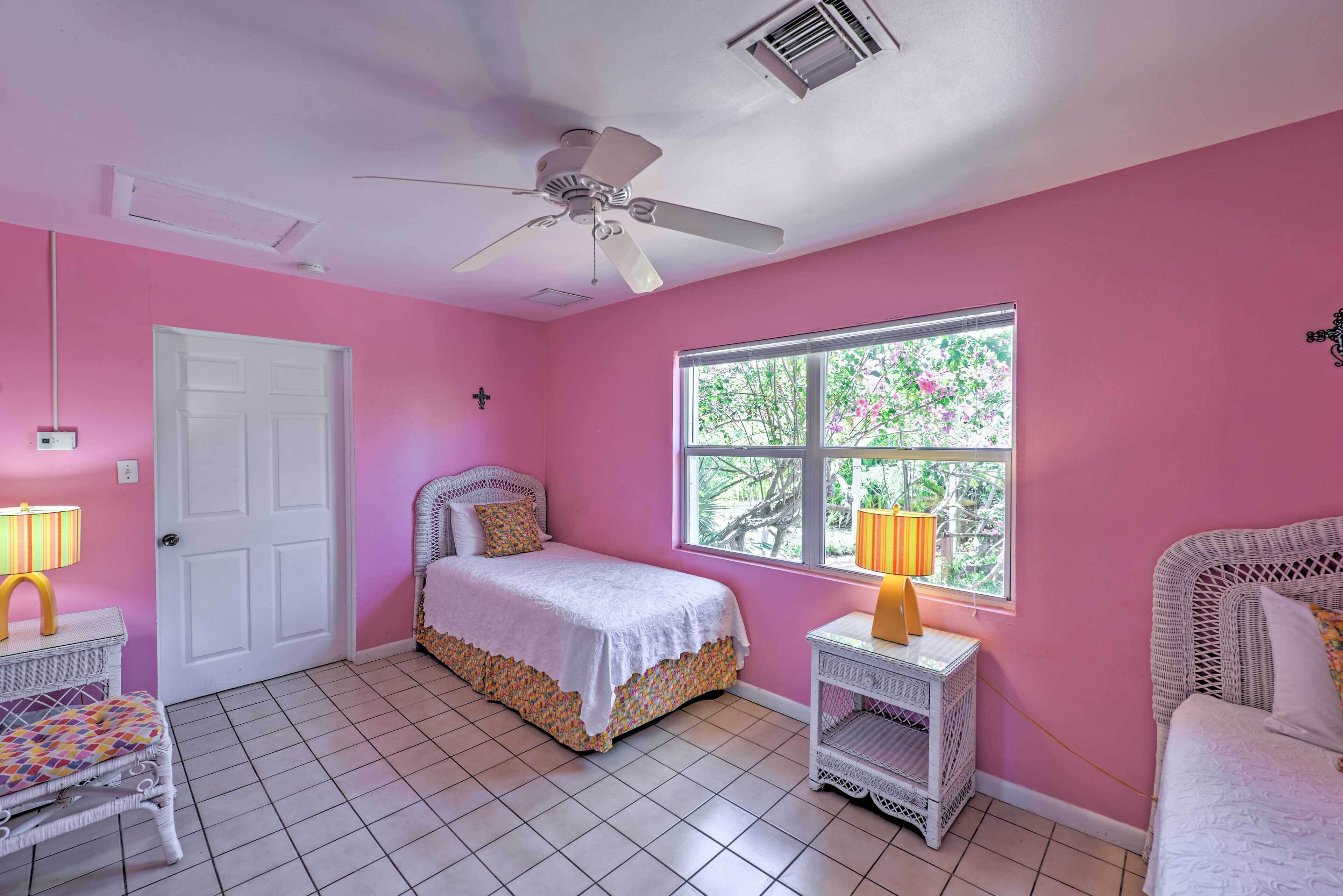 A pink-themed bedroom with a bed, a window with a view, a ceiling fan, and a white door.
