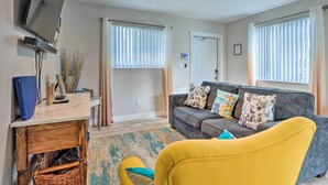 Un salon avec un canapé, un fauteuil jaune, un téléviseur fixé au mur et un ventilateur de plafond.