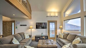 A living room with a large sectional sofa, a leather ottoman, and two floor lamps. There is a flat-screen TV mounted on the wall and a window with a view of the outside. - Logan Road Lookout, Lincoln City Home w/ Game Room (Lincoln City)