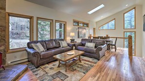 A spacious living room with two leather sofas, a wooden coffee table, and large windows offering views of a wooded area. - Creekside Mtn House w/ Deck: 8 Mi to Idaho Springs (Dumont)
