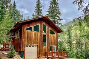 A wooden cabin with a garage, surrounded by tall trees and a mountainous backdrop. - Creekside Mtn House w/ Deck: 8 Mi to Idaho Springs (Dumont)