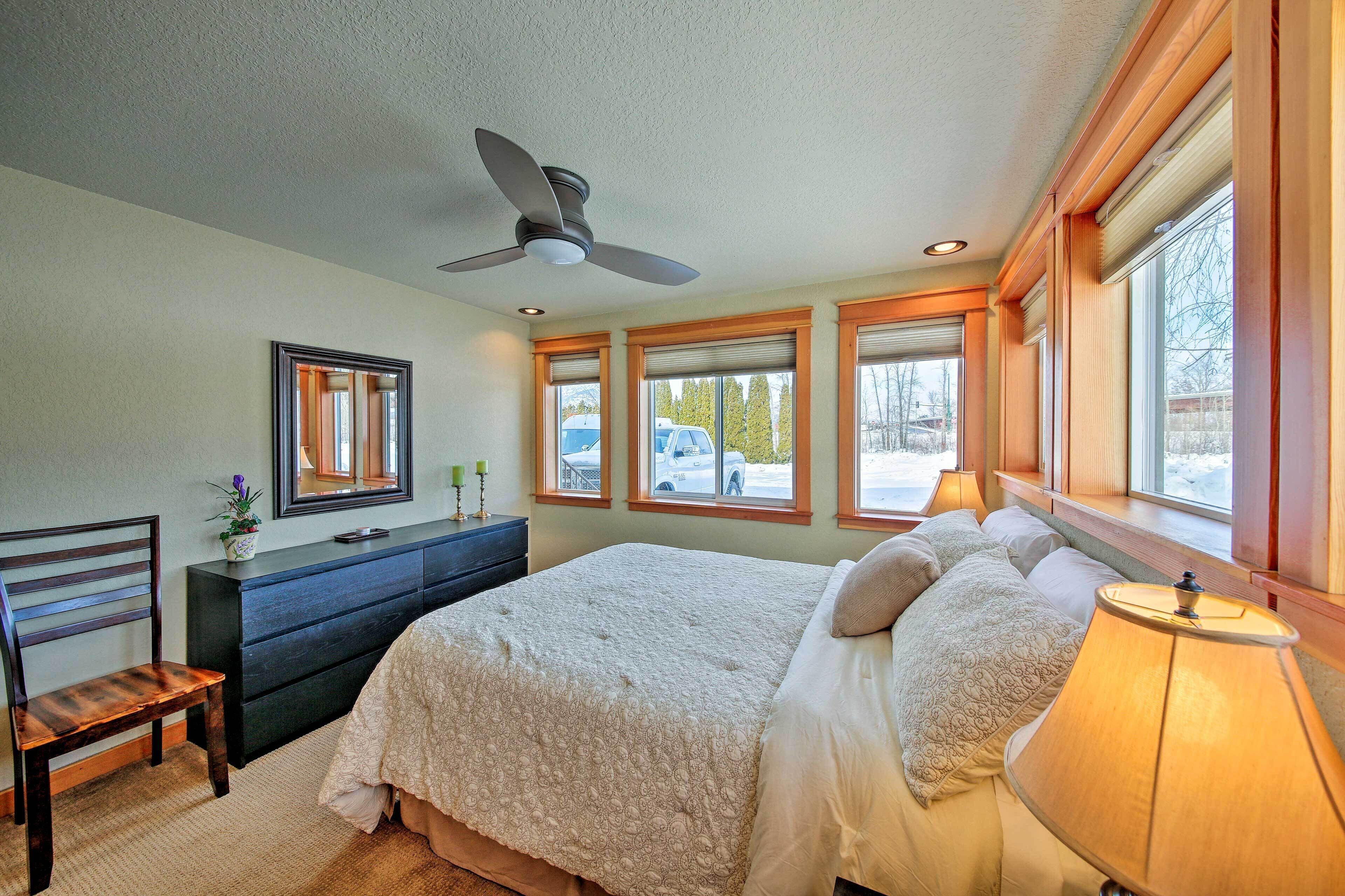 Un dormitorio con cama, escritorio, silla, ventilador de techo y ventanas amplias.