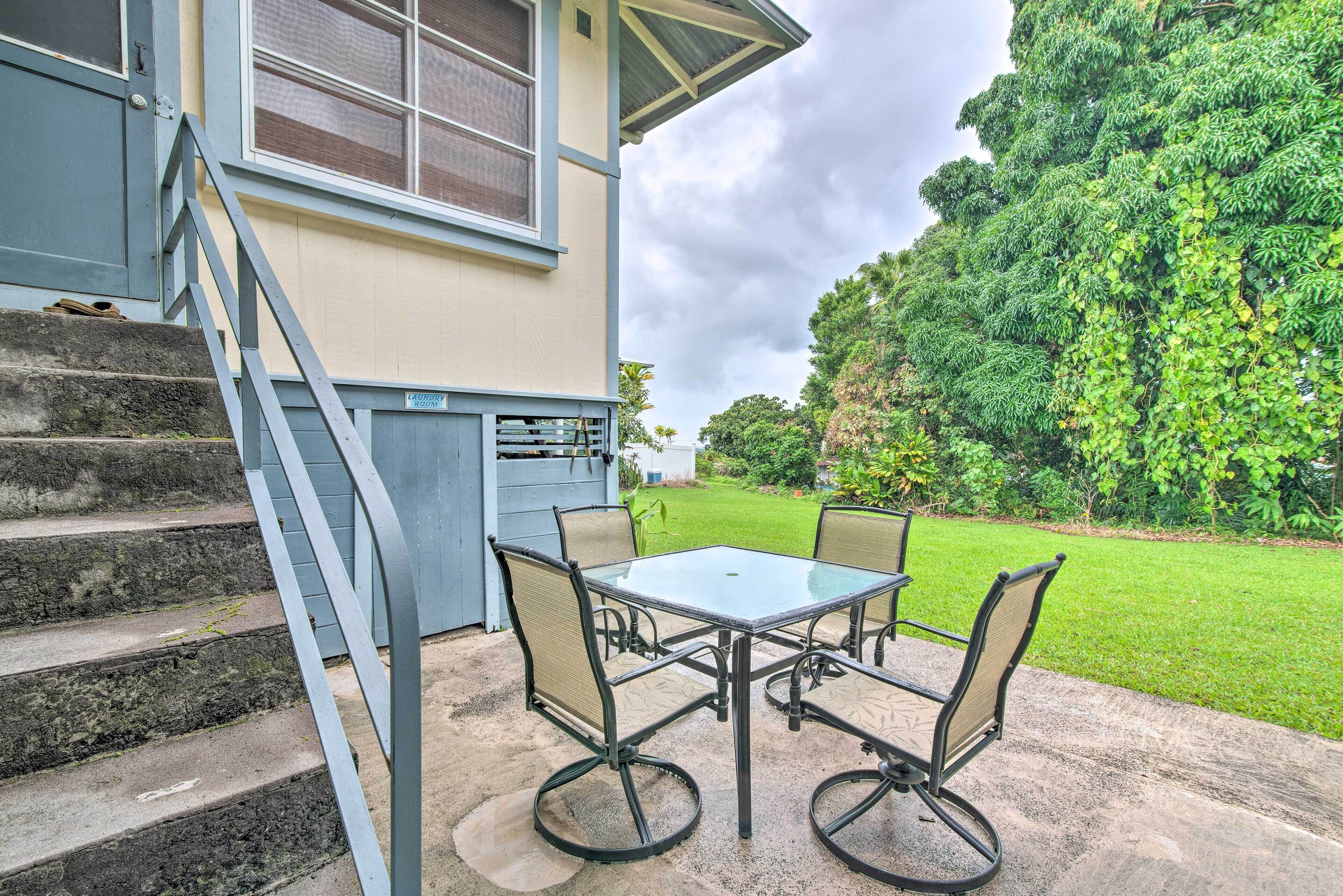 A patio with a glass table and chairs, a set of stairs leading to a door, and a lush green garden in the background.