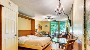 A bedroom with a large bed, a glass table, wooden chairs, a ceiling fan, and a chandelier. - Kailua-kona Islander Studio: Lanai, Community Pool (Kailua-Kona)