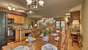 A dining area with a wooden table set for four, surrounded by wooden chairs. A chandelier hangs above, and a potted orchid is on the table.