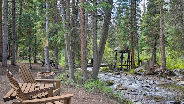 A forested campsite with wooden chairs and a picnic table, a small wooden structure, and a stream.