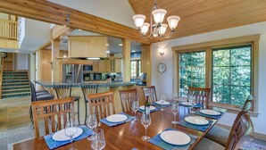 A spacious dining area with a wooden table set for six, surrounded by chairs. A kitchen is visible in the background, and there is a large window providing a view of trees. - Mountain Home ~ 5 Mi to Winter Park Resort! (Winter Park)