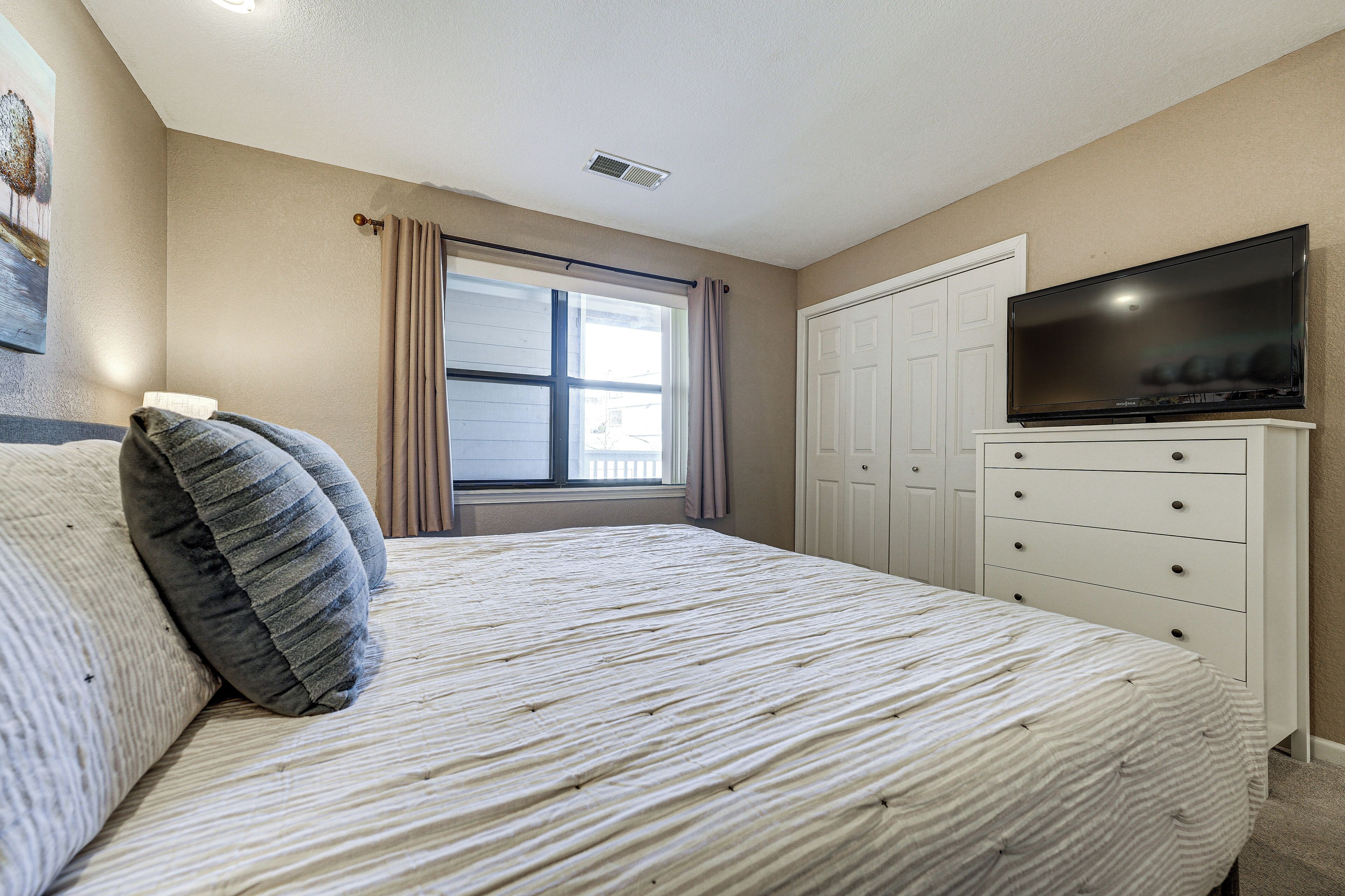 A bedroom with a bed, a television on a dresser, a window with curtains, and a door.