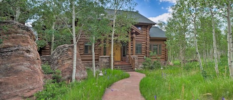 A log cabin with a stone pathway leading to the entrance, surrounded by lush greenery and trees.