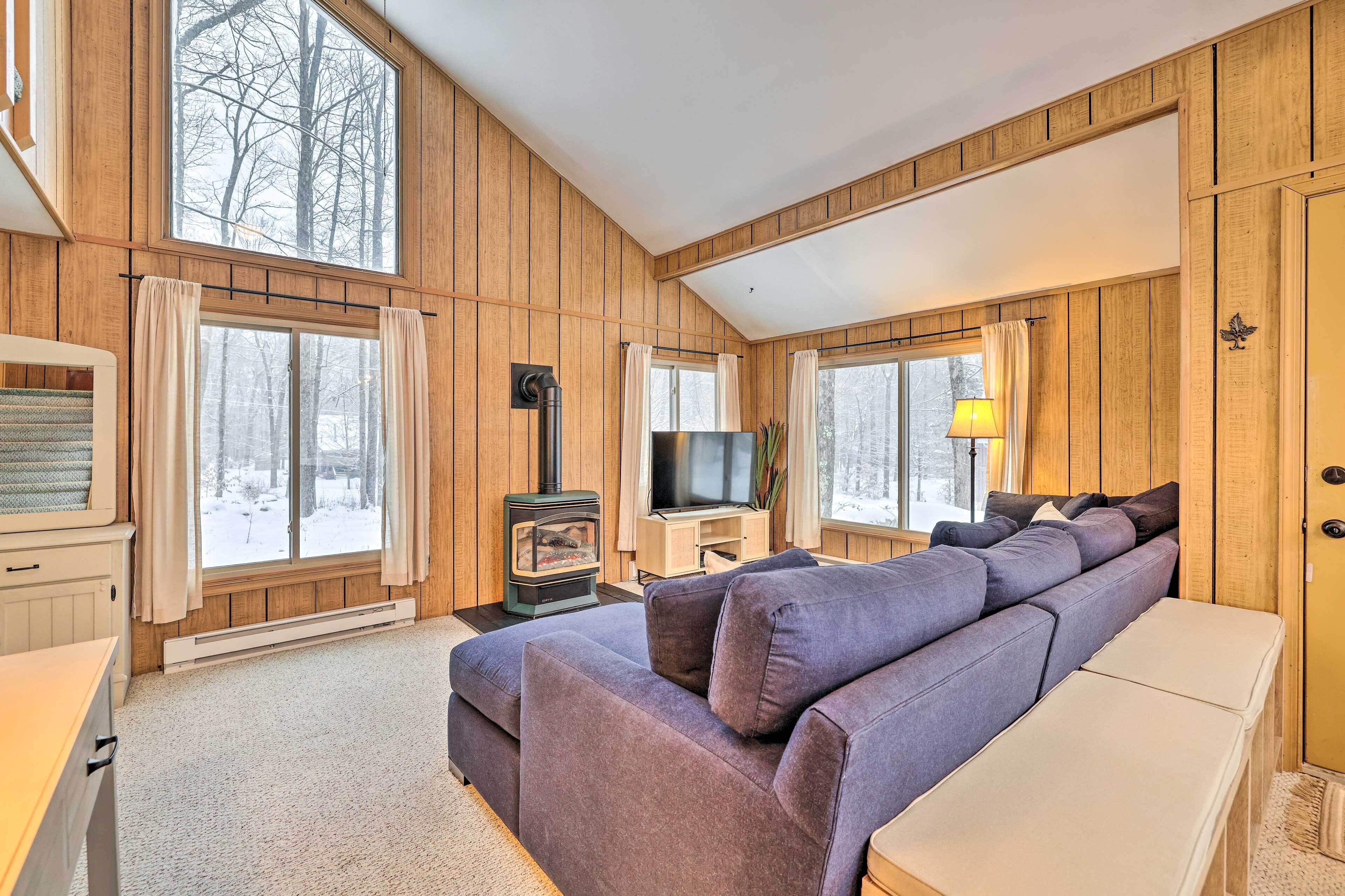 Ein gemütliches Blockhauszimmer mit großem Fenster, Kamin, Sofa und Fernseher.